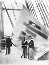 Two soldiers with rifles and one man in a sailor suit standing on a ship deck in front of a large flag.