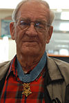 Photograph of an older white man wearing glasses and a medal hanging from a blue ribbon around his neck