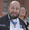 Head and shoulders of a bald man with dark beard, with a medal hanging from a blue ribbon around his neck.