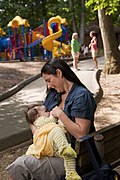 Woman breastfeeds infant on bench at children's playground.