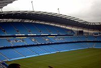 Roughly the same camera position shows grass up to the blue seats of the stands. The stand is now split into three levels of permanent seating.