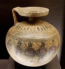 Oplite su un aryballos globulare. Gli aryballos appaiono spesso appesi con una piccola cinghia al polso dell'atleta nelle scene di palestra. Erano anche un bene di sepoltura popolare e sarebbero stati un'offerta votiva adatta in un santuario di eroi (Payne 1931, 290E).