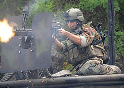 Un membro dell'equipaggio della US Navy Boat Team che spara con una mitragliatrice pesante. Nota l'armatura difensiva e il cinturone delle munizioni; cartucce esaurite che cadono. Equipaggio in tenuta mimetica, equipaggiamento radio, telecamera sul casco, mitragliatrice sopra la spalla.