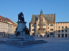 Im Vordergrund eine im Schatten liegende auf einem steinernen Podest stehende Statue. Am Podest sind zwei weitere Statuen platziert. Dahinter steht neben anderen, von der Sonne beschienen Häusern zentral ein mit Wappen verziertes fachwerkartiges Gebäude mit einem zentralen Turm und einer Uhr.