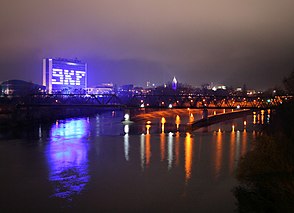 Im Vordergrund Wasser. Dahinter eine rechts orange beleuchtete Eisenbahnbrücke, im Hintergrund eine Skyline mit einem links herausstechenden blau beleuchteten rechteckigen Haus mit der Beschriftung „SKF“.