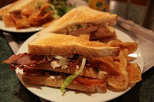Um sanduíche torrado de dois decks contendo bacon, tomate, alface e ovo servido com batatas fritas em um prato de porcelana branca. Este sanduíche muito provavelmente tem maionese espalhada sobre ele em vez de ou em adição à manteiga. Um sanduíche semelhante é mostrado no fundo, mas este tem salada em vez de batatas fritas. Ambos os pratos estão em uma bandeja, que está em um balcão de mármore cinza ou mármore com efeito de balcão.