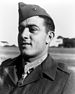 Head of a young white man in a plain dark jacket with a star-shaped medal hanging from a ribbon around his neck. His garrison cap is tilted to the side and has a single round pin on its side.