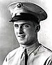 Head and shoulders of a white man wearing a light colored shirt and tie and a peaked cap with a dark visor.