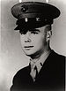 Head and shoulders of a young white man wearing a dark peaked cap and a dark military jacket over a shirt and tie. On the collar of the shirt is a bar-shaped pin.