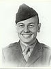 Head and shoulders of a young white man with a broad smile. He is wearing a garrison cap tilted over his right ear and a plain military jacket on top of a shirt and tie.