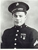 Head and shoulders of a young white man wearing a peaked cap and a military jacket with large shiny buttons, two chevrons on the upper sleeves, and two medals and two ribbon bars on the left breast.