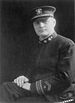 A seated man, his hands folded and resting on his knee, in military uniform with stripes on his sleeve near the wrist, oak leaf and anchor emblems on his collar, and a peaked cap.