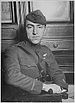 Head and torso of a man turned in his chair, his right arm laying stiffly on the arm rest in front of him. He is wearing a military uniform with a strap diagonally across the chest, a winged emblem on his left breast, and a garrison cap.