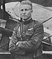 Head and torso of a white man with short, light, hair and a stern look on his face. His arms are crossed and he is wearing a military uniform with a strap across the chest, a high stiff collar, and a winged badge on his left breast. Behind him can be seen part of a biplane.