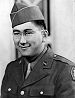 Head and shoulders of a smiling young man with dimples and round wire-framed glasses wearing a garrison cap and a military jacket over a shirt and tie.