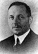 Head of a middle-aged white man with thinning hair, wearing a dark suit coat, white shirt, and dark tie.