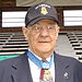 Head and shoulders of an elderly white man wearing glasses and a baseball cap with an image of a star-shaped medal and the words "Medal of Honor recipient". An actual star-shaped medal hangs from a light blue ribbon around his neck, over his dark suit coat and patterned tie.