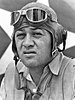 Head of a squinting man wearing a shirt unbottoned at the collar and a cloth aviator's cap with headphones built into the ear flaps, an unbuckled chin strap, and goggles pushed up onto his forehead.