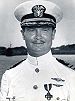 Head and shoulders of a white man with a thin mustache wearing a white peaked cap, its black visor decorated with oak leaves, and a white military jacket with dark shoulder boards and one medal and two winged pins on the left breast.