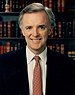 A color image of Kerrey from the waist up wearing a suit and tie. He is smiling at the camera and there is a book shelf with books behind him.