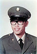 Head and shoulders of a young man wearing a peaked cap, black thick-rimmed glasses, and a military jacket with a round pin on each lapel over a shirt and tie.