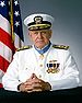 Head and torso of an elderly white man, in front of an American flag, with his hands folded and resting on a table in front of him. He is wearing a white jacket with a row of medals hanging from ribbons and a winged pin on his left breast, gold shoulderboards, and a star-shaped medal hanging from a light blue ribbon around his neck. His peaked cap is white with a black visor decorated with gold leaves, a gold band around the brim, and an eagle-anchors-and-shield emblem on the front.