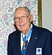 Elderly white man wearing a suit, tie, and glasses, with a medal hanging from a ribbon around his neck.