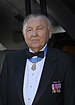 Head and shoulders of an elderly white man with a round face, wearing a tuxedo with military medals pinned to the chest and one medal hanging from a light blue ribbon around his neck.