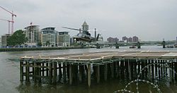 Letoun Sikorsky S-76A přistává na heliportu London Heliport na molu na řece Temži. V pozadí je železniční most Battersea