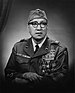 Head and torso of a man sitting with his arms folded on his lap, wearing a garrison cap, horn-rimmed glasses, and a military jacket. The jacket's left breast is completely covered in ribbon bars and medals.