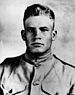 Head and shoulders of a square-jawed young man with short, light, hair, which stands straight up in the front. He is wearing a military jacket with breast pockets and a row of buttons down the center.