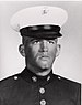 Head and shoulders of a young white man with a cleft chin wearing a dark military jacket and a white peaked cap with a black visor.