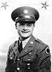 Head and shoulders of a smiling young white man wearing a peaked cap, wire-framed glasses, and, over a shirt and tie, a jacket adorned with pins on the lapels, stripes and a round badge on the shoulder, and a whistle on a chain hanging from a shoulder button.